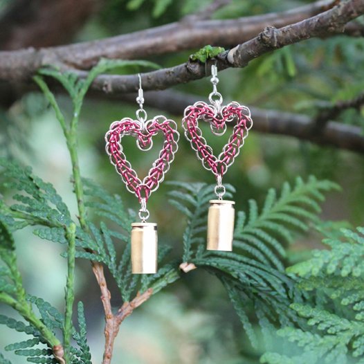 Handmade chainmail heart earrings in red and silver hanging from a cedar branch. The earrings feature upcycled 9mm brass bullet casings dangling from the bottom, symbolizing spiritual alchemy and transformation.